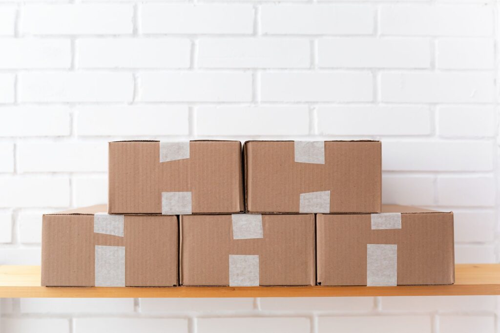 A neat stack of cardboard boxes taped and placed on a wooden shelf against a white brick wall.