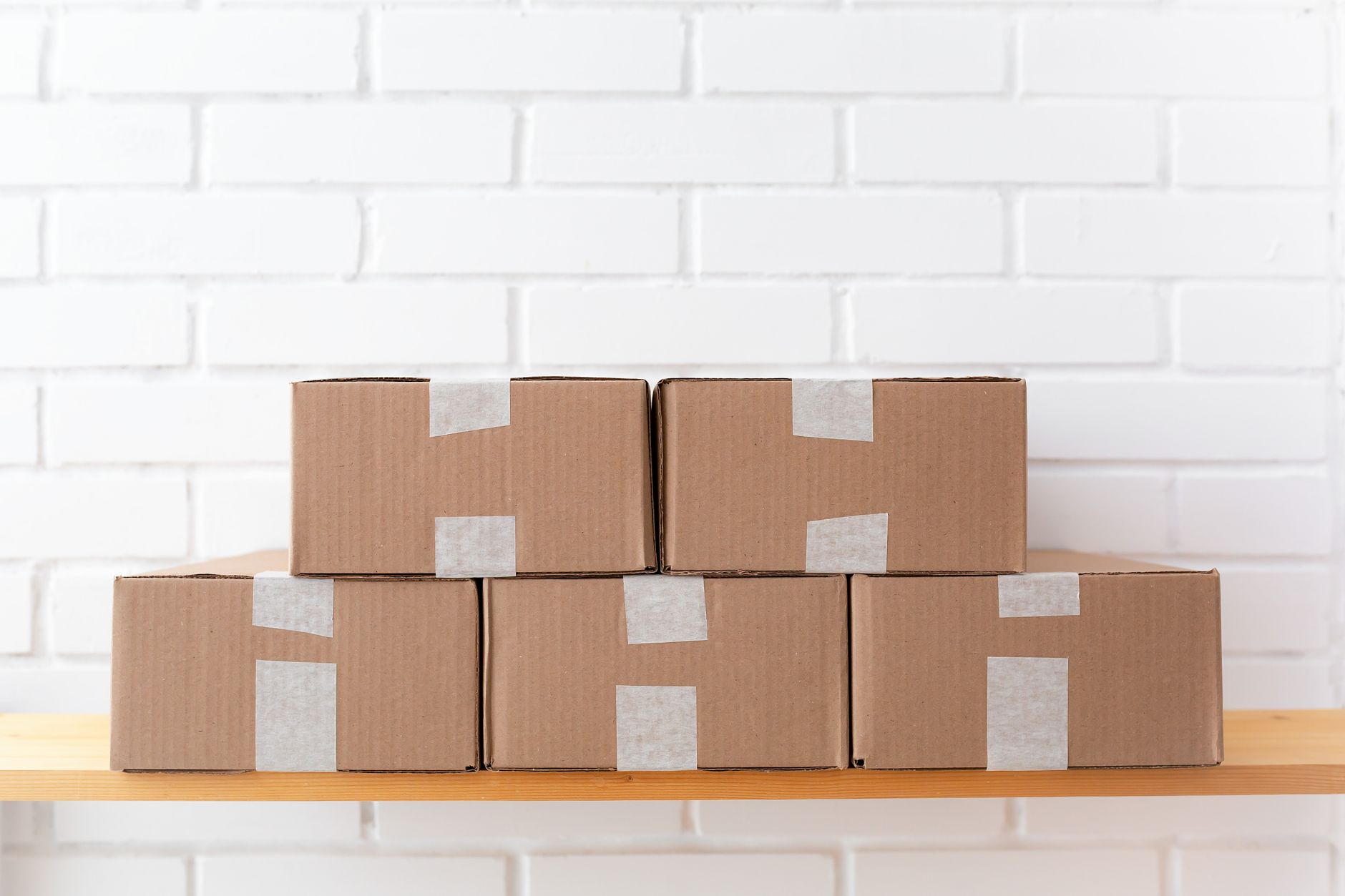 A neat stack of cardboard boxes taped and placed on a wooden shelf against a white brick wall.