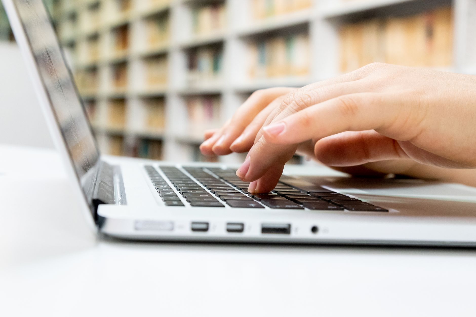 Close-up of hands typing on a laptop keyboard in a library setting.